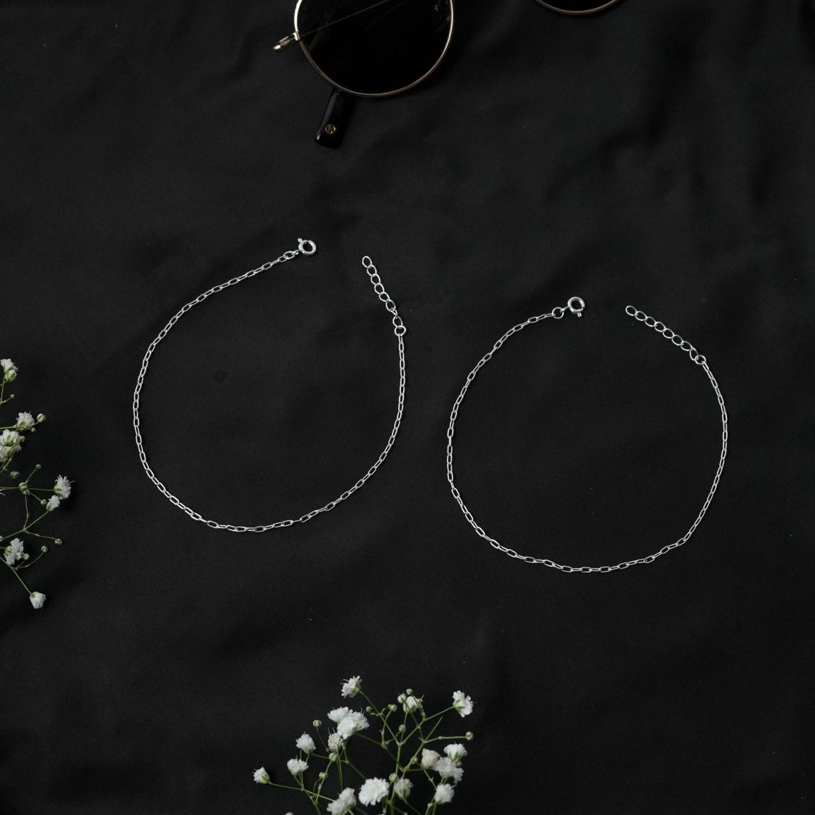 Two silver chain anklet on a dark fabric background with small white flowers.