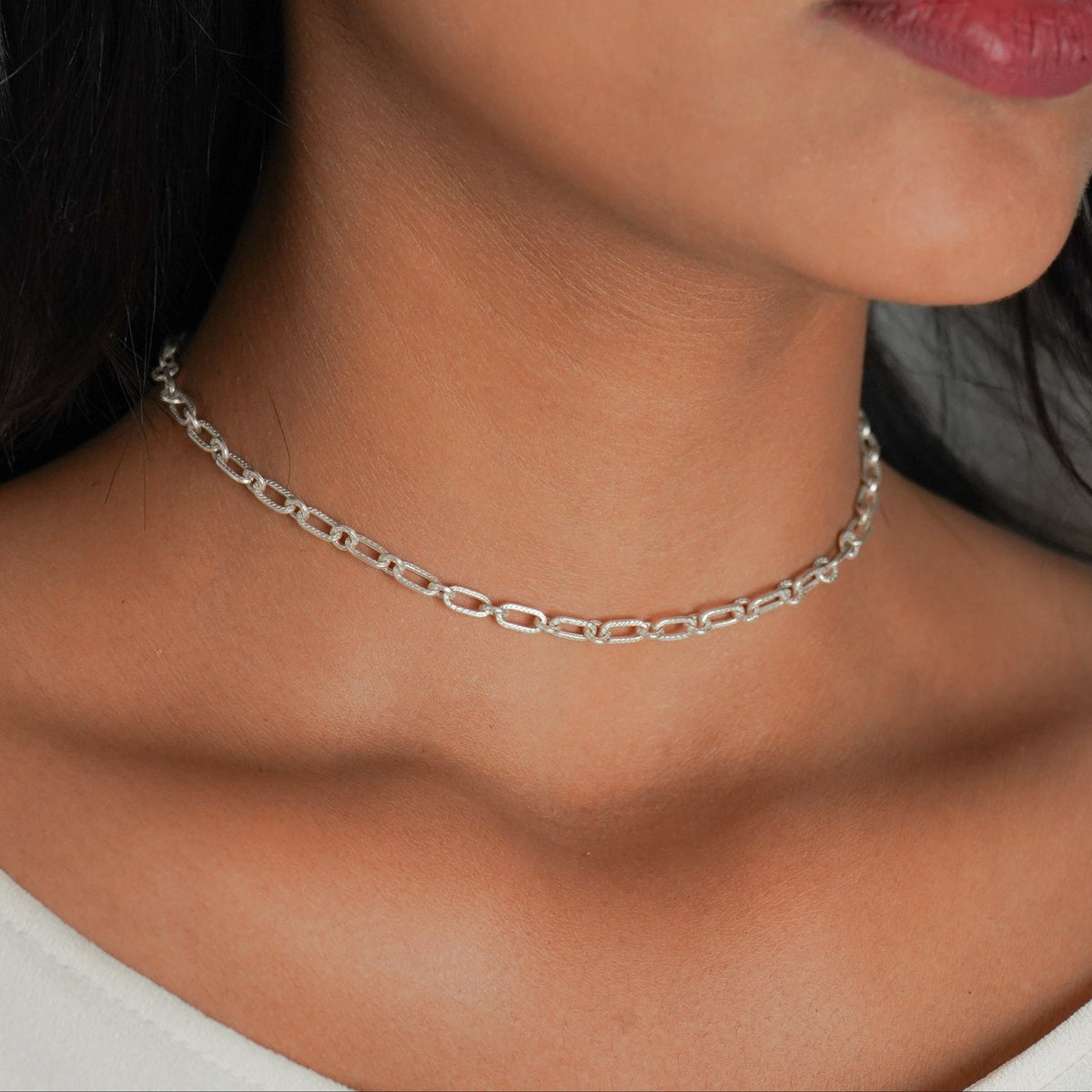 A side view of a woman wearing a delicate silver chain choker necklace with a textured, openwork pattern, paired with a white top.