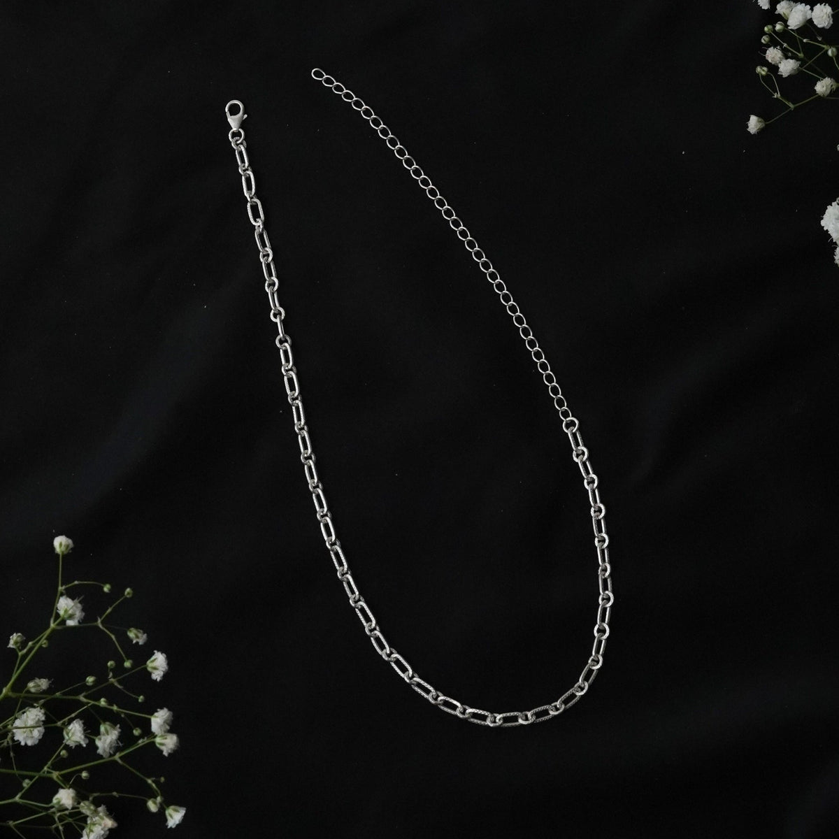 A 925 silver paperclip chain necklace with a lobster clasp, showing on a black fabric background with small white baby's breath flowers.