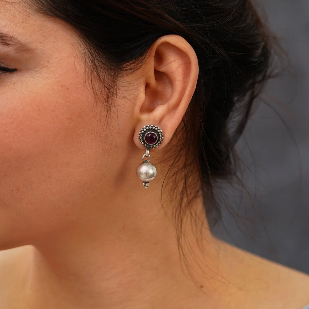 Close-up of a woman wearing a 925 silver drop earring with a red stone hanging with a round silver ball on a neutral background.