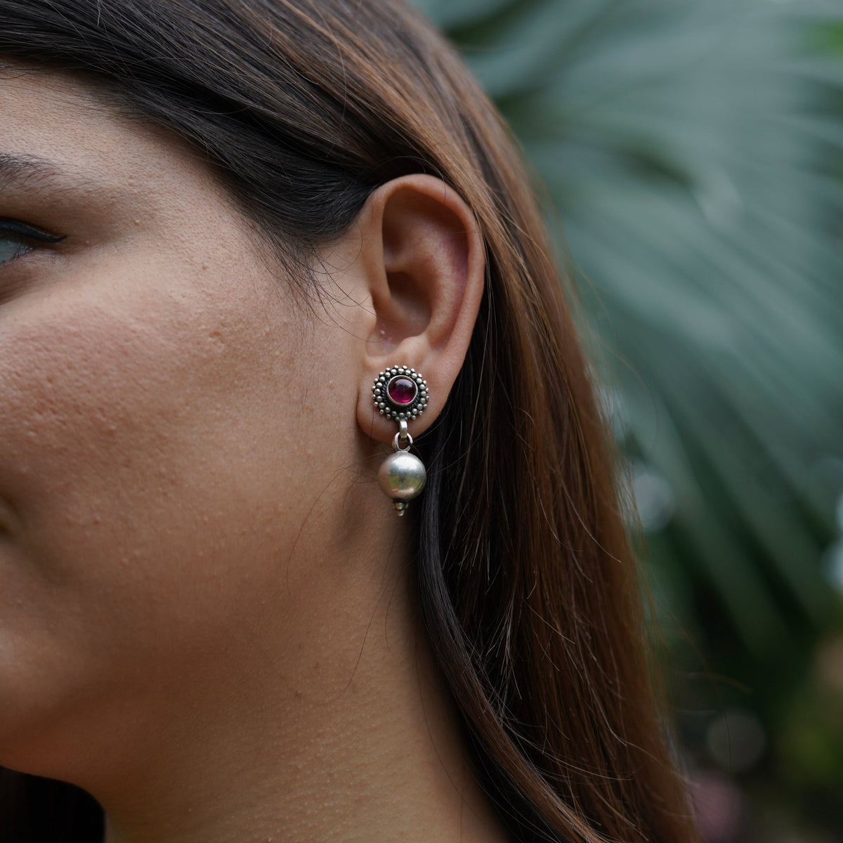Close-up of a women wearing a 925 silver drop earring featuring a red stone link with a silver ball attach with a green leafy background.
