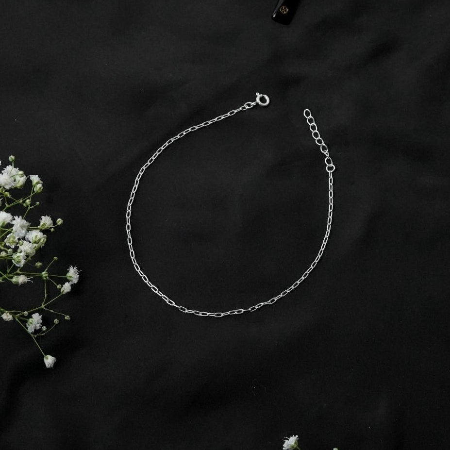 A delicate silver chain anklet with a lobster clasp and extender, laid out on a black fabric background with small white flowers as decorative props.