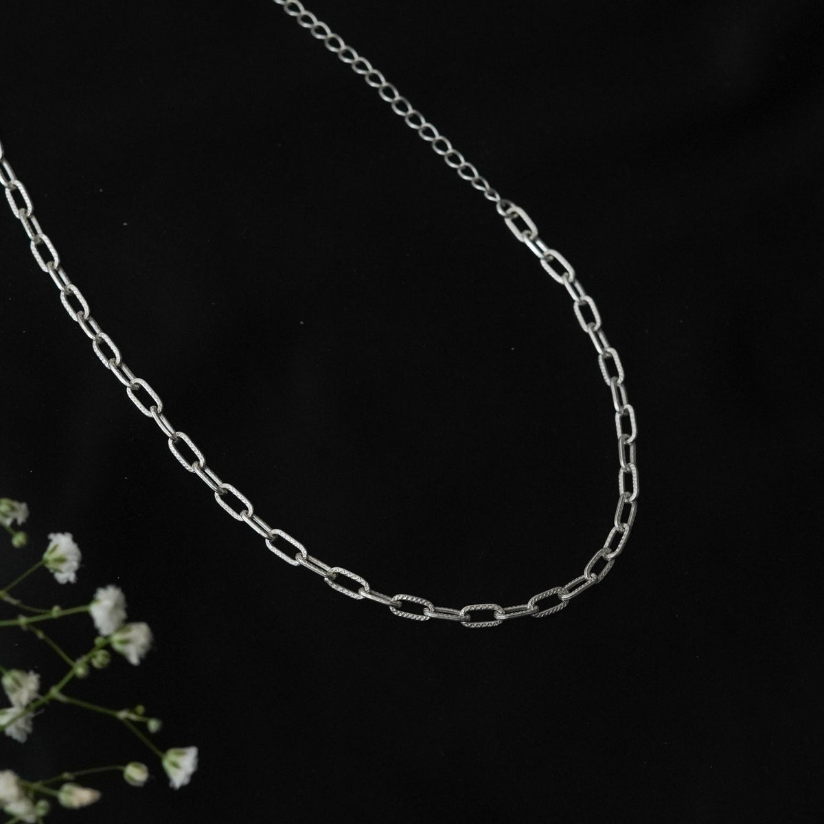 A 925 sterling silver paperclip chain necklace displayed against a black background with white breath flowers.