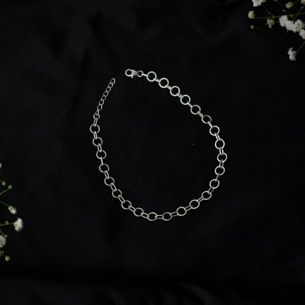 A 925 silver chain anklet with oval links and a lobster clasp, displayed on a black fabric background with small white flowers in the corners.